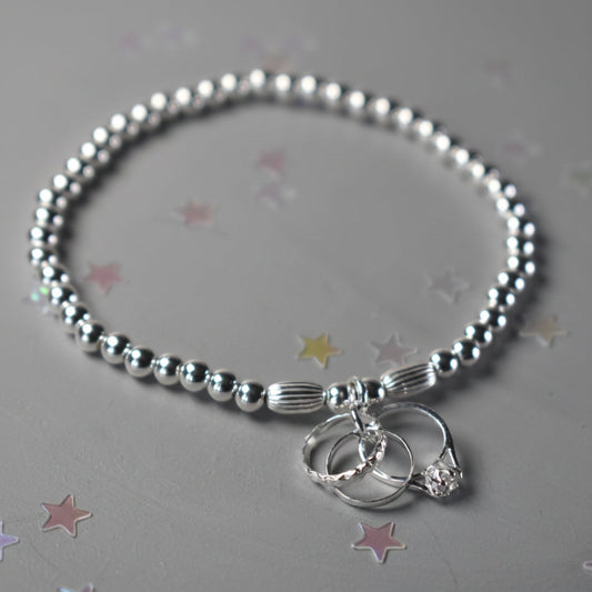 An elasticated sterling silver beaded bracelet with 4mm beads finished with a sterling silver charm of three rings symbolising engagement , wedding and eternity rings. 